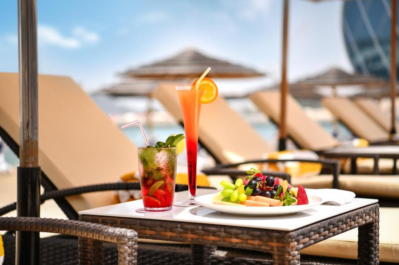 Al Raha Beach Resort & Spa Abu Dhabi Exterior photo The photo shows a table set outdoors, featuring two colorful cocktails and a plate of food. One cocktail is a vibrant red drink garnished with mint, while the other is a layered orange beverage with an orange slice on the rim. The plate of food inclu