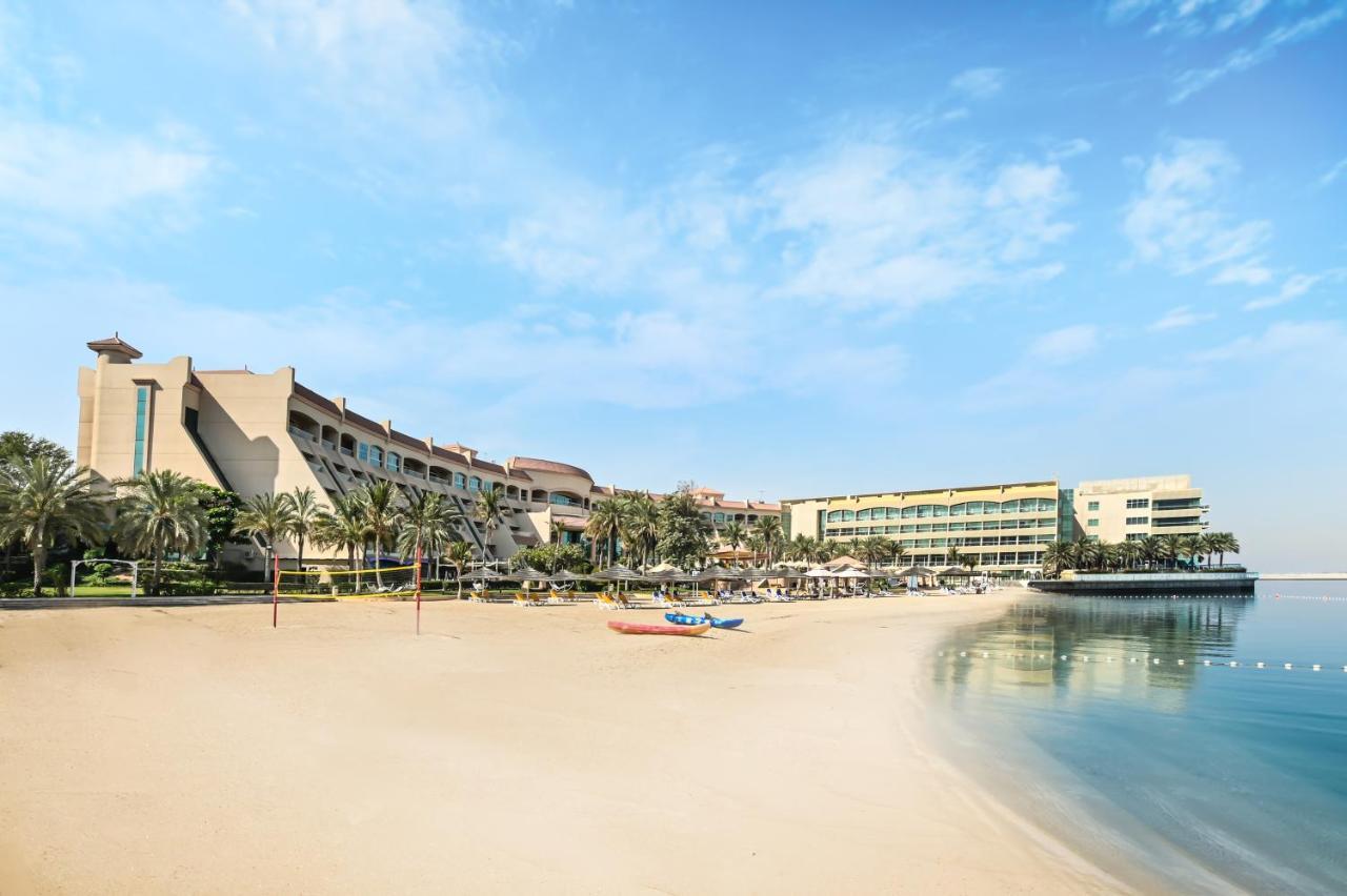 Al Raha Beach Resort & Spa Abu Dhabi Exterior photo The photo depicts a serene beach scene with a clear blue sky. In the foreground, there is a sandy beach that gently slopes down to calm water. On the left side, several palm trees provide a tropical atmosphere. In the background, there are two large 