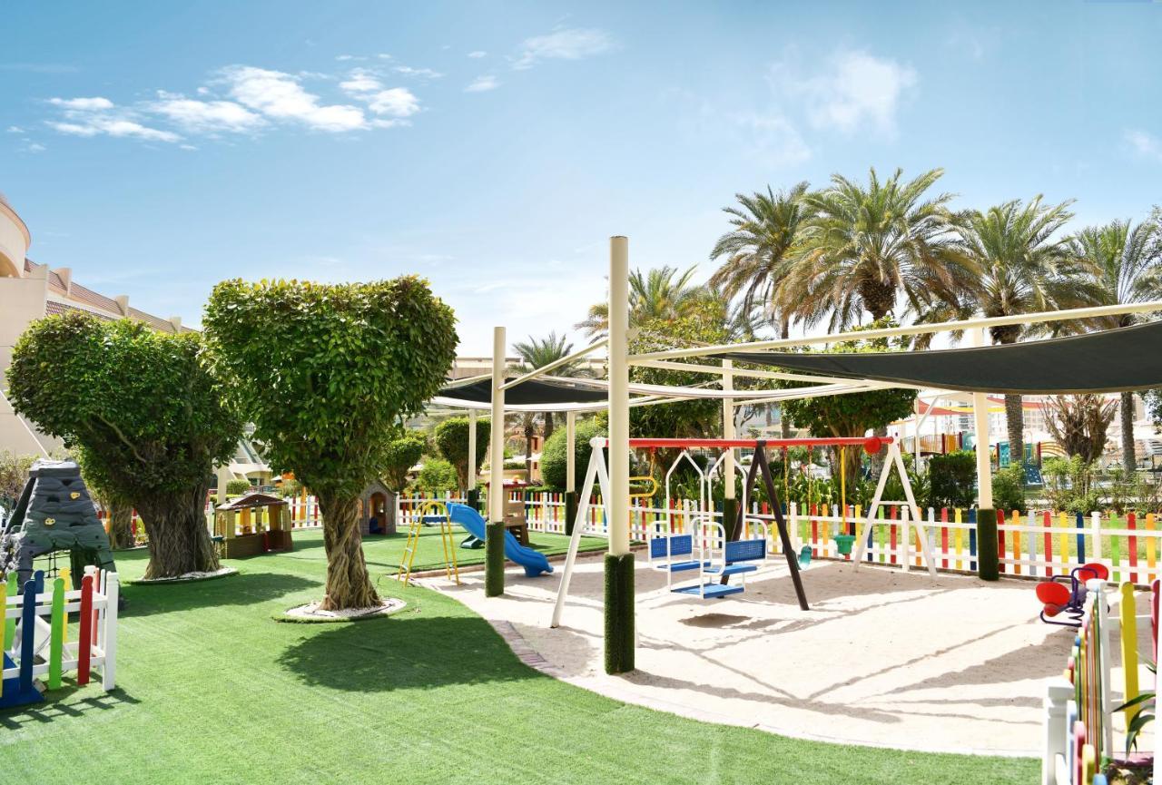 Al Raha Beach Resort & Spa Abu Dhabi Exterior photo The photo shows a brightly lit outdoor playground surrounded by palm trees. The playground features a variety of play equipment, including swings, a slide, and a climbing structure. The ground is covered in lush green artificial grass, providing a so