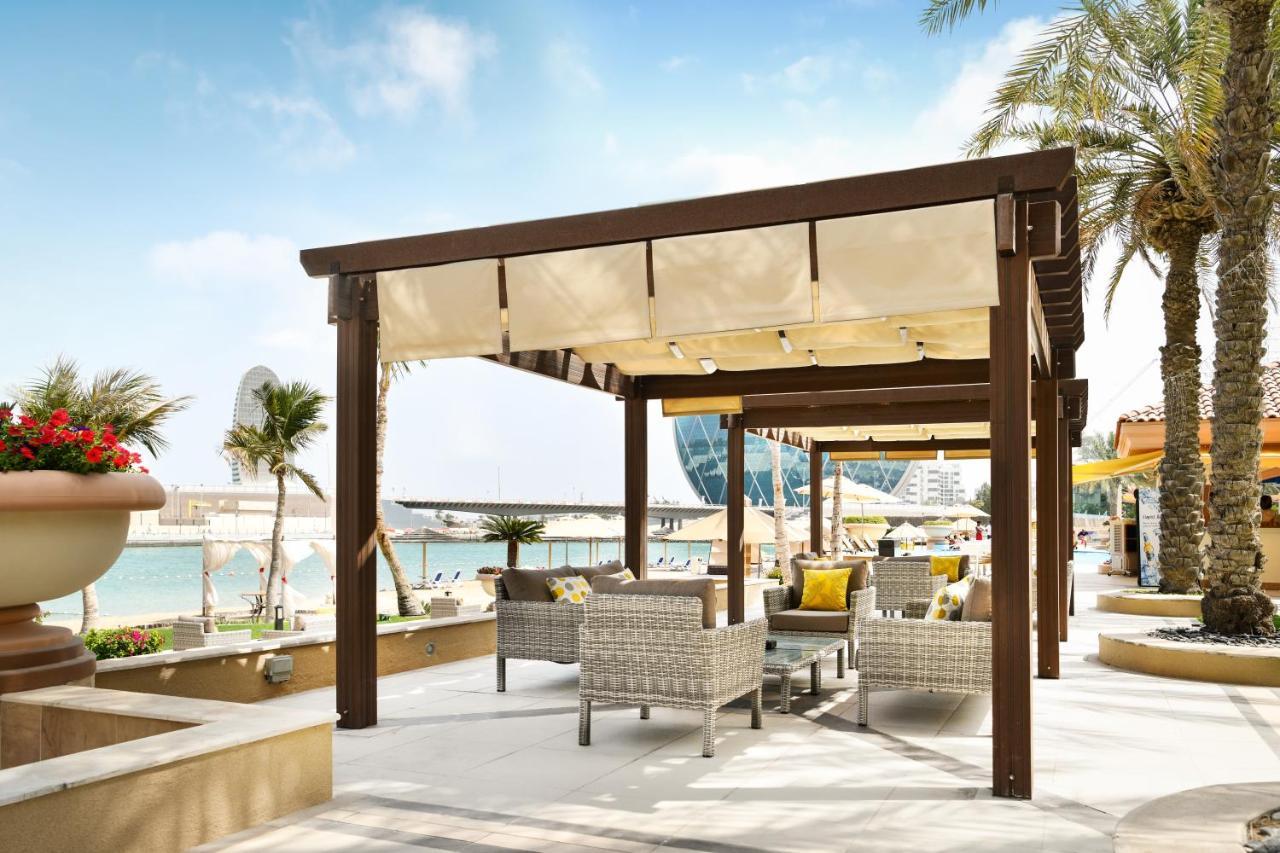 Al Raha Beach Resort & Spa Abu Dhabi Exterior photo The photo shows a stylish outdoor seating area by a beach. There is a wooden pergola covered with a light fabric shade, providing shelter to a set of comfortable wicker chairs and tables. The area is adorned with palm trees, and in the background, th