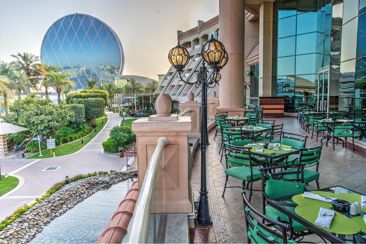 Al Raha Beach Resort & Spa Abu Dhabi Exterior photo The photo depicts an outdoor dining area with green tables and chairs arranged on a patio. In the background, there is a large, distinctive building with a striking blue dome, surrounded by well-maintained landscaping, including trees and plants. The
