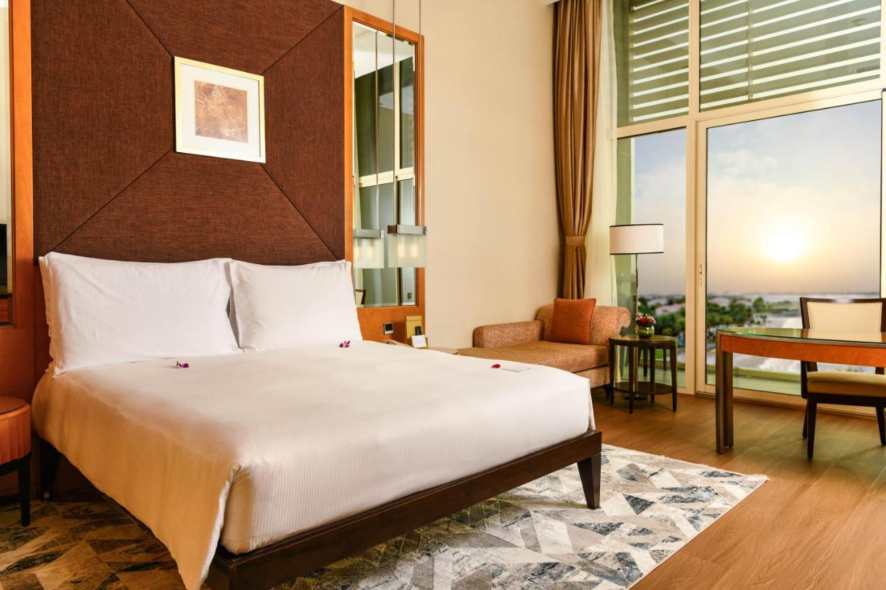 Al Raha Beach Resort & Spa Abu Dhabi Exterior photo The photo shows a stylish hotel room featuring a large bed with white bedding, positioned against a textured wall. There’s a small seating area with a chair and a table, and a window with curtains that allows natural light to fill the space. The room