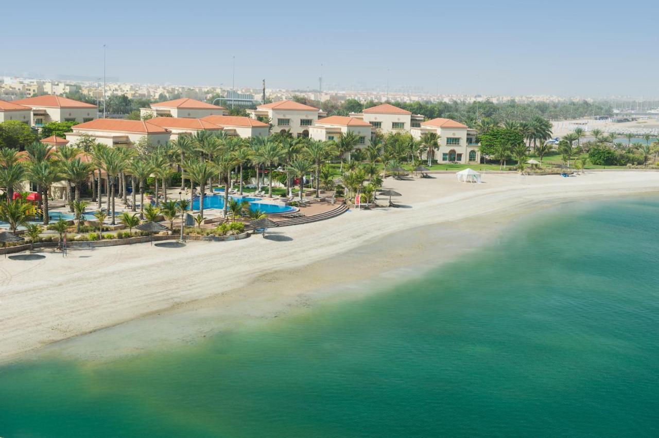 Al Raha Beach Resort & Spa Abu Dhabi Exterior photo The photo depicts a serene beach scene with a clear turquoise sea and a sandy shoreline. In the background, there are several buildings featuring a Mediterranean architectural style with red-tiled roofs. The area is adorned with palm trees and outdoo