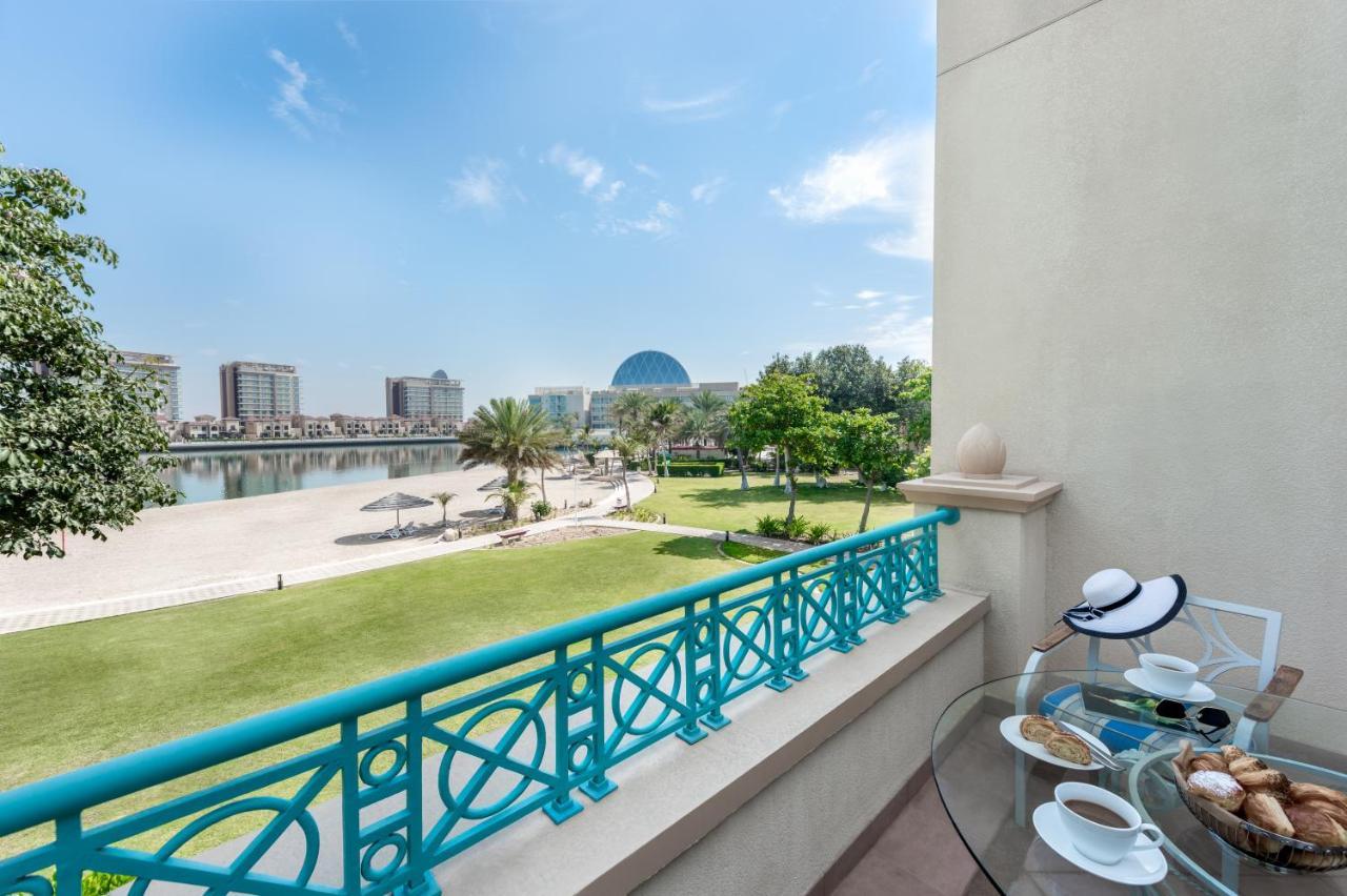 Al Raha Beach Resort & Spa Abu Dhabi Exterior photo The photo shows a balcony view overlooking a scenic landscape. In the foreground, there is a glass table set with snacks or pastries and two cups. Beyond the balcony railing, there is a well-maintained green area with palm trees and a pathway. The ba