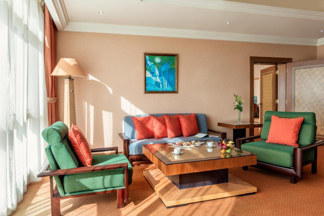 Al Raha Beach Resort & Spa Abu Dhabi Exterior photo The photo shows a cozy living room with a warm ambiance. There are two sofas: one in green with red cushions and another in blue with red cushions, arranged around a wooden coffee table. On the table, there are some fruits and a small decorative item