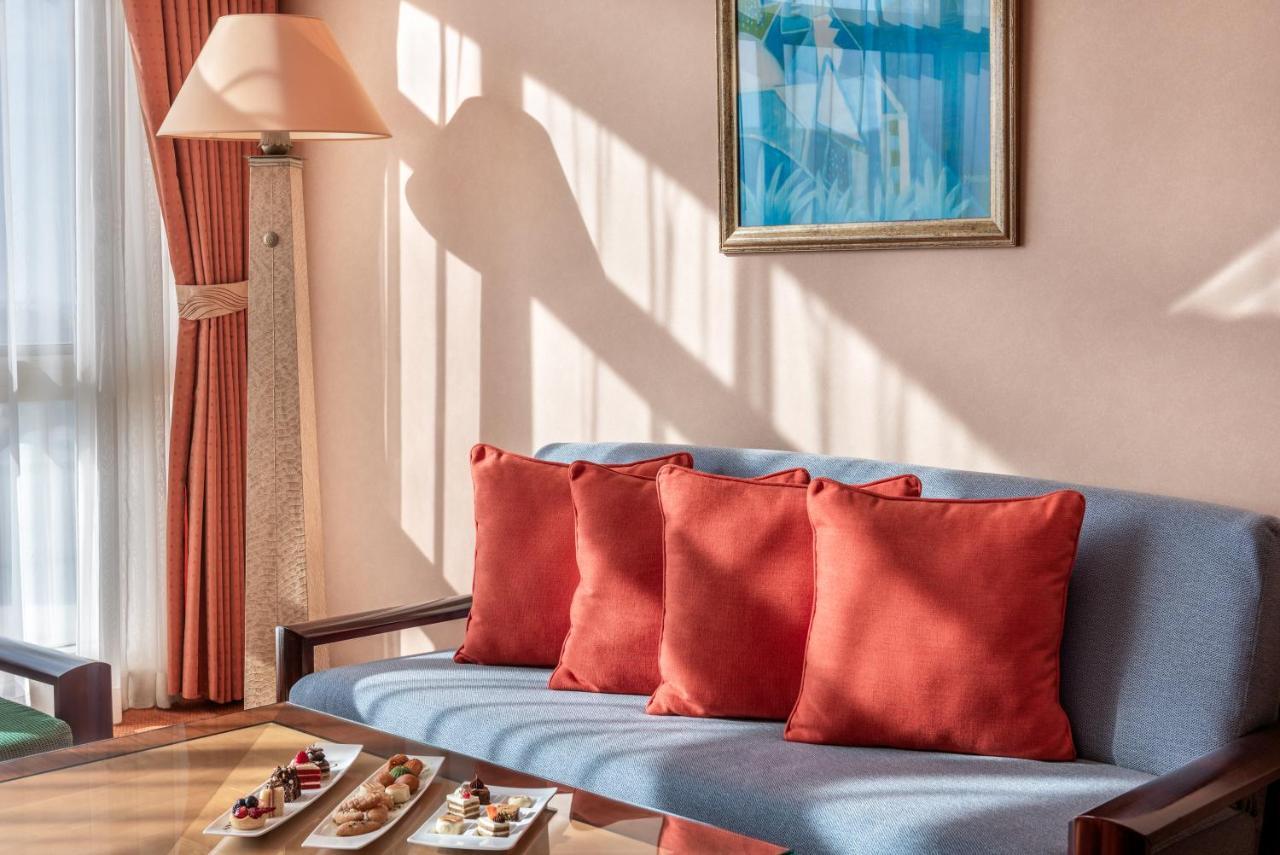 Al Raha Beach Resort & Spa Abu Dhabi Exterior photo The photo shows a cozy interior setting, likely a sitting area. It features a light blue couch with four red cushions. In front of the couch, there is a glass table displaying an assortment of snacks or small dishes. The room has soft lighting, with 