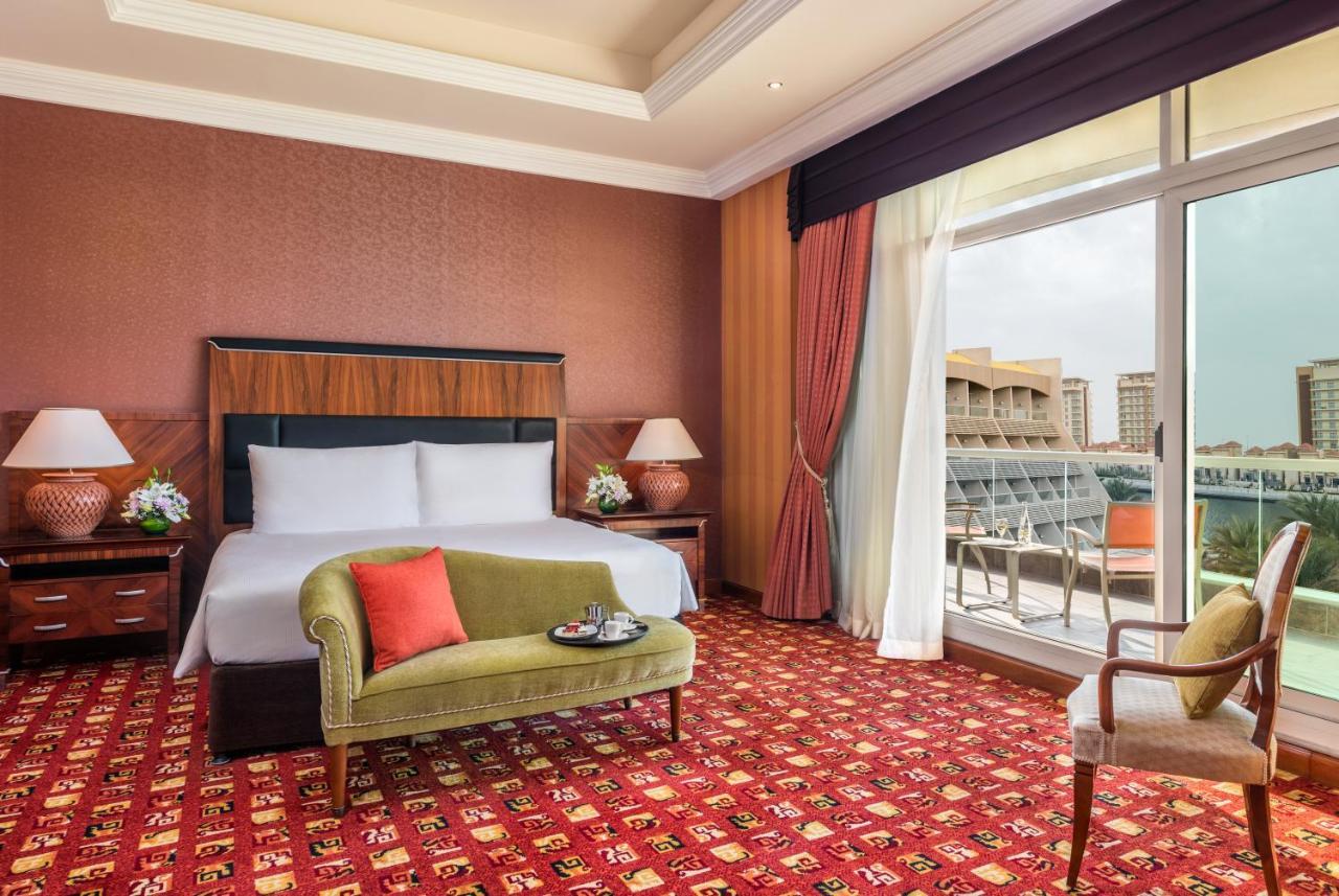 Al Raha Beach Resort & Spa Abu Dhabi Exterior photo The photo shows a hotel room with an elegant and modern design. It features a large bed with white linens and a dark wooden headboard. On either side of the bed are matching bedside lamps. There is a green armchair with a red pillow positioned near t