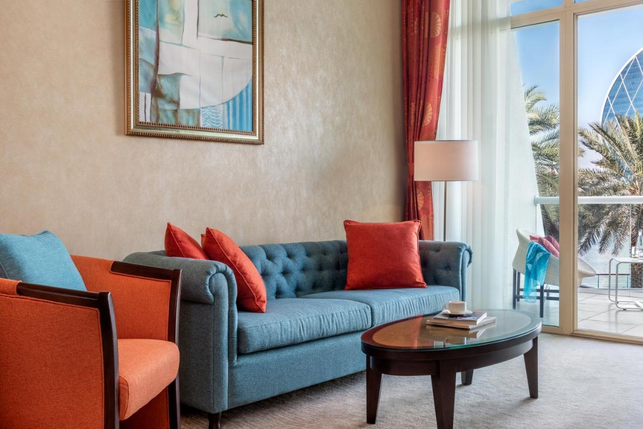Al Raha Beach Resort & Spa Abu Dhabi Exterior photo The photo shows a well-decorated living room. There is a plush blue couch with orange accent pillows and a cozy orange armchair nearby. A round coffee table sits in front of the couch, and above it, there is a colorful painting hanging on the wall. L