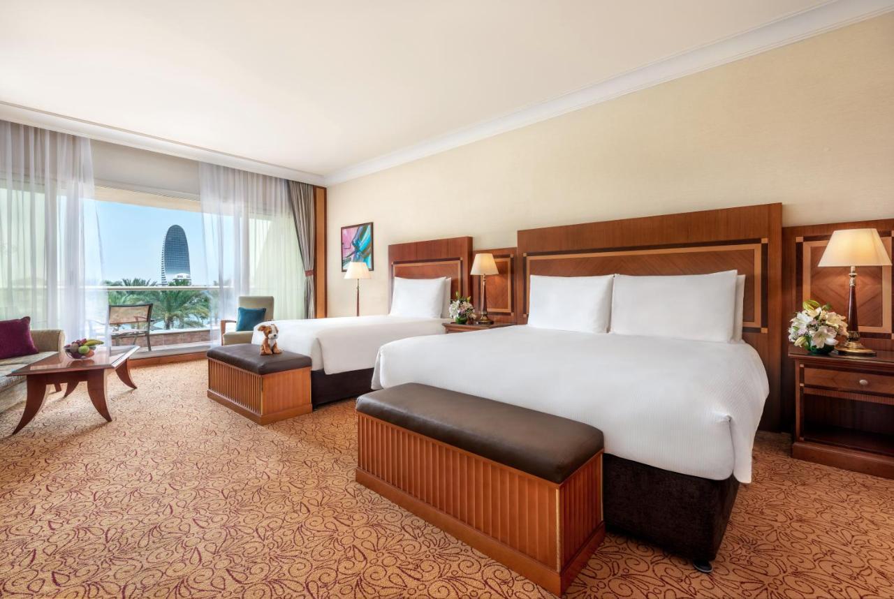 Al Raha Beach Resort & Spa Abu Dhabi Exterior photo The photo shows a spacious hotel room featuring two large beds with white linens and dark brown upholstered headboards. There are matching benches at the foot of each bed. The room is decorated with a warm color palette and has a carpeted floor. To o