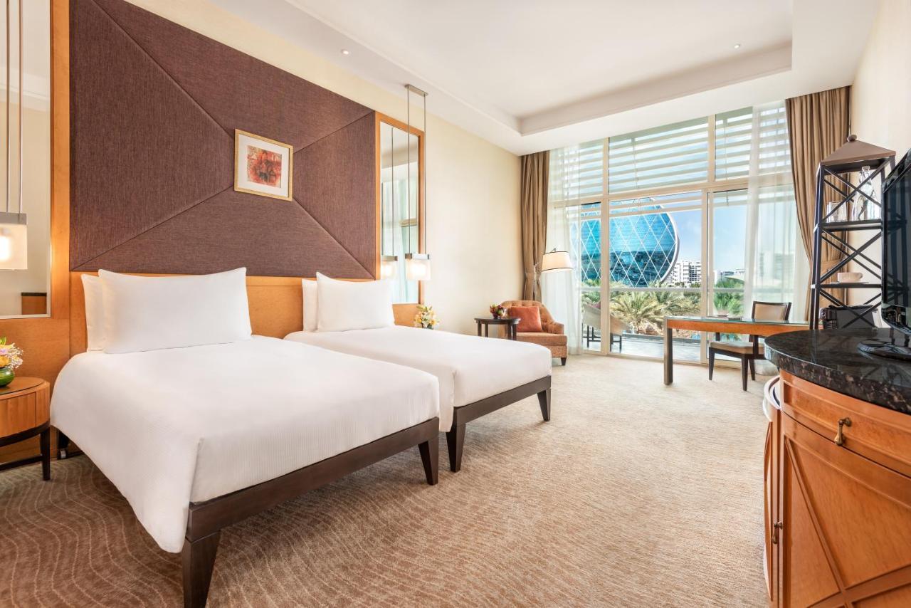 Al Raha Beach Resort & Spa Abu Dhabi Exterior photo The photo shows a well-decorated hotel room featuring two single beds with white linens. The room has a modern aesthetic, with a large window letting in natural light and offering a view of a circular structure outside, possibly a landmark or artisti