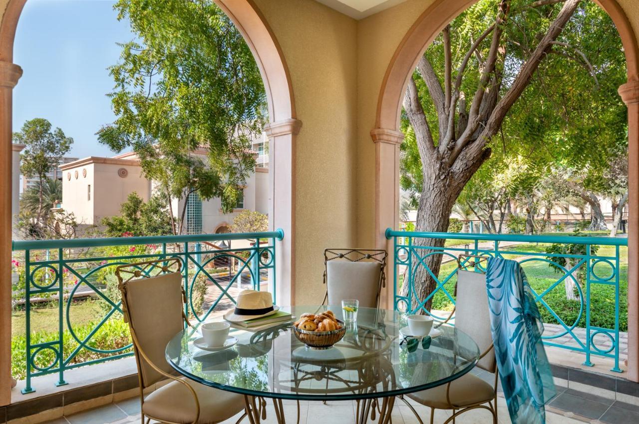 Al Raha Beach Resort & Spa Abu Dhabi Exterior photo The photo shows a serene outdoor balcony setting. There is a circular glass table with a bowl of fruit or snacks in the center, alongside a glass and a small plate. Surrounding the table are four elegant chairs. The railing is turquoise, framing a vi