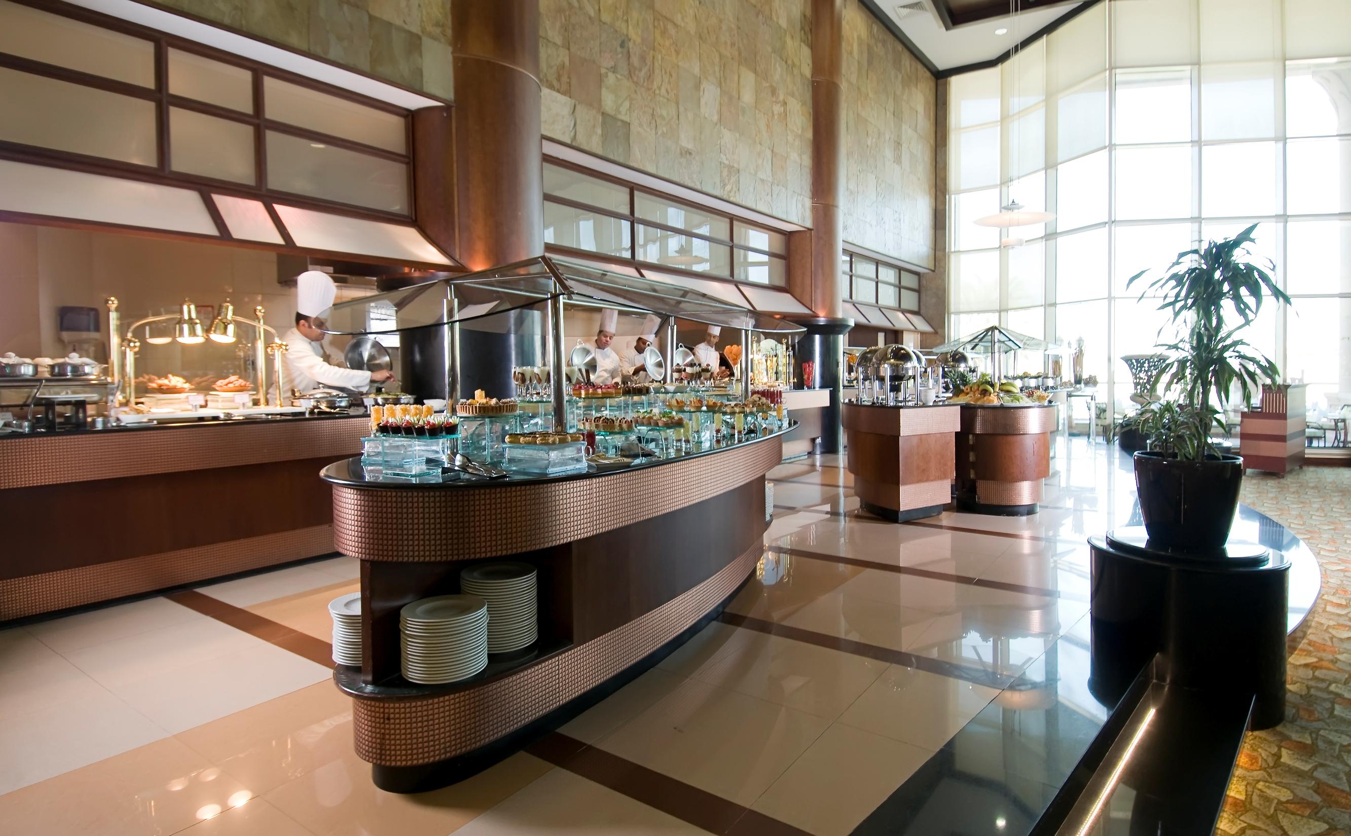 Al Raha Beach Resort & Spa Abu Dhabi Exterior photo The photo shows a spacious buffet area in a restaurant or hotel. There are multiple food stations with glass displays showcasing a variety of dishes. Chefs can be seen in the background, preparing food. The decor features large windows allowing natur