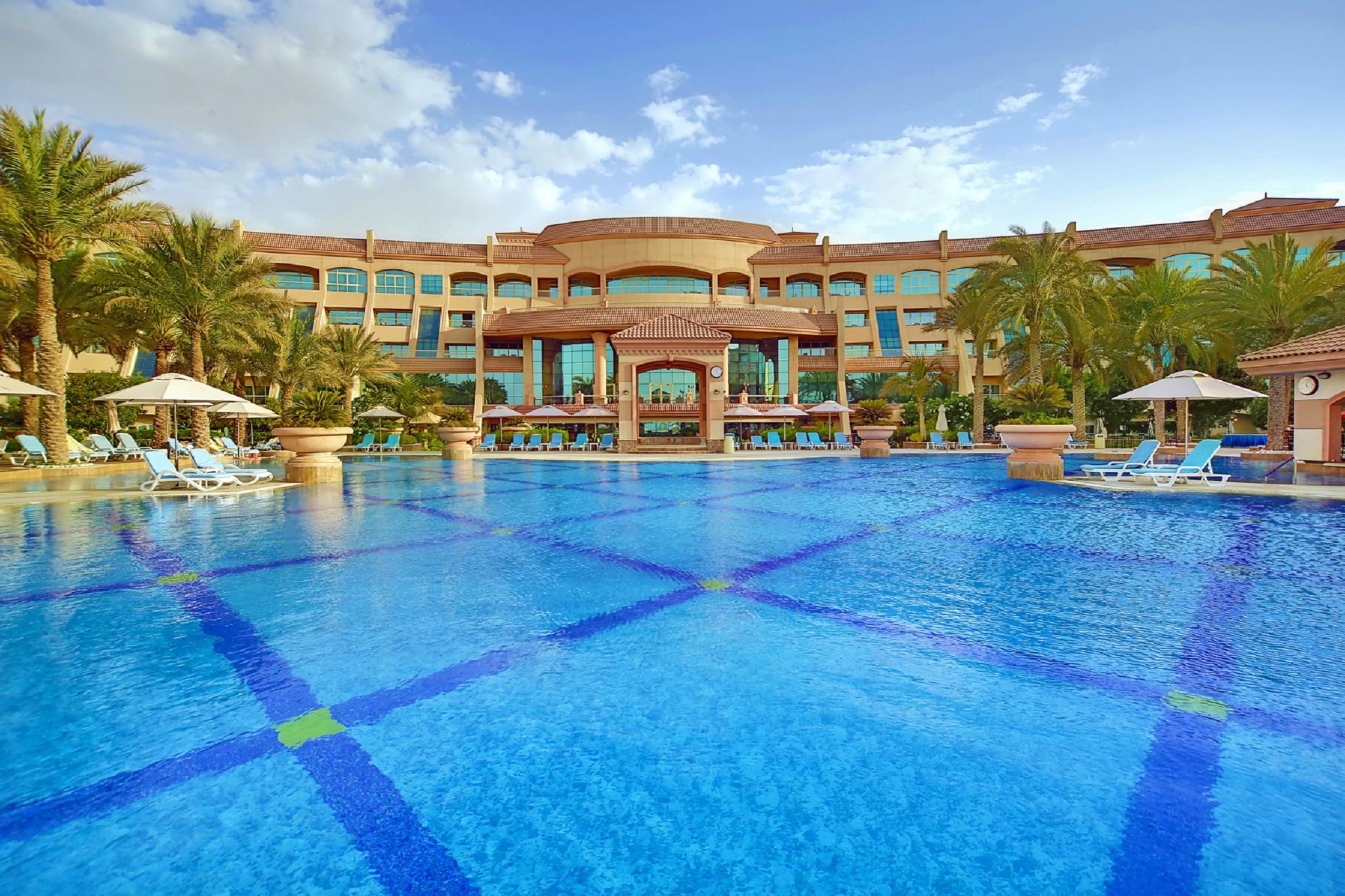Al Raha Beach Resort & Spa Abu Dhabi Facilities photo The photo features a luxurious resort with a large swimming pool in the foreground. The pool is crystal clear and reflects the sky, bordered by lounge chairs and palm trees. In the background, the resort building showcases modern architecture with la
