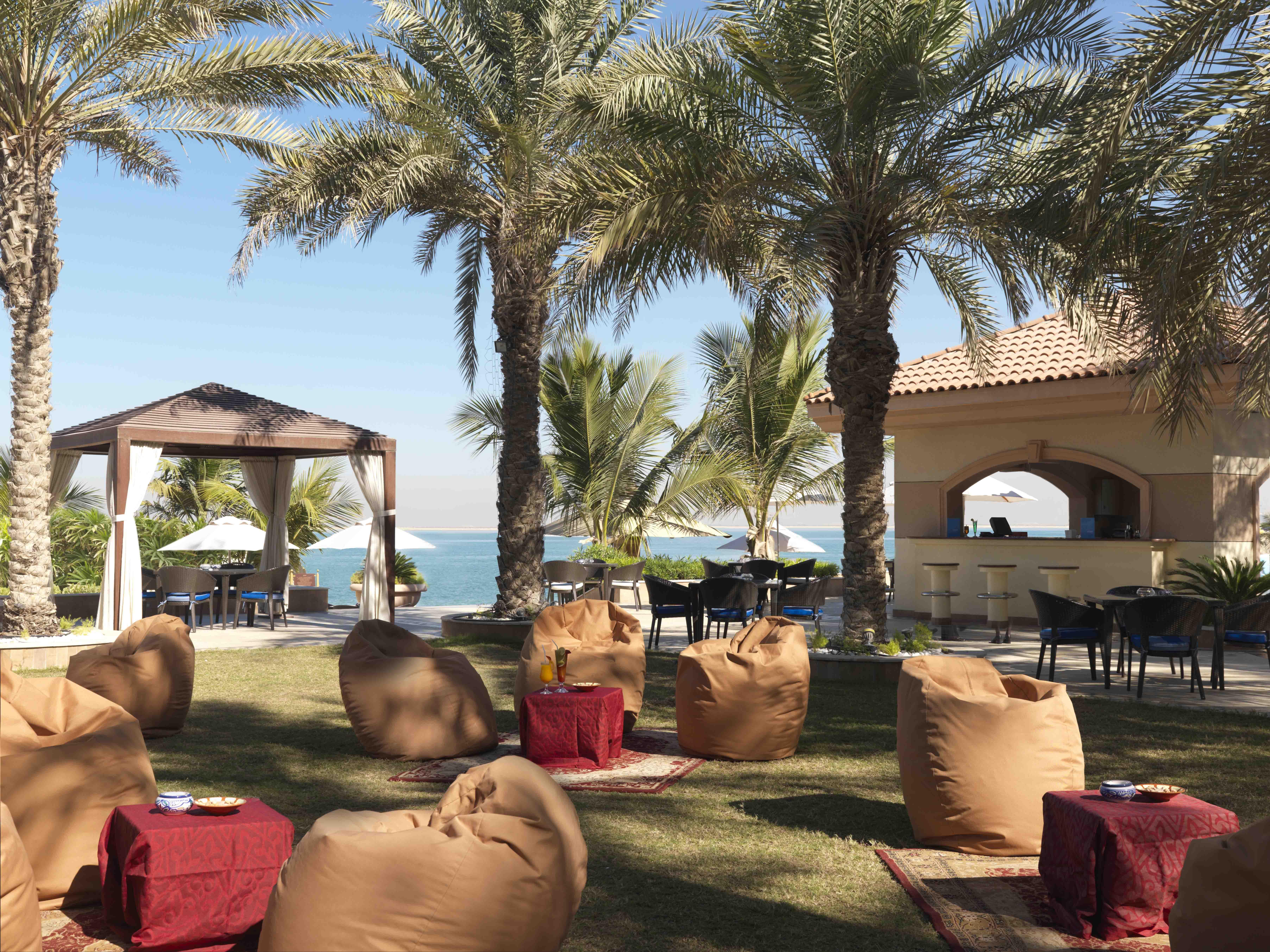 Al Raha Beach Resort & Spa Abu Dhabi Restaurant photo The photo depicts a beautiful outdoor setting with several elements of relaxation and leisure. In the foreground, there are large, comfortable bean bag chairs arranged on a grassy area, accompanied by low tables. Surrounding this seating area are tal