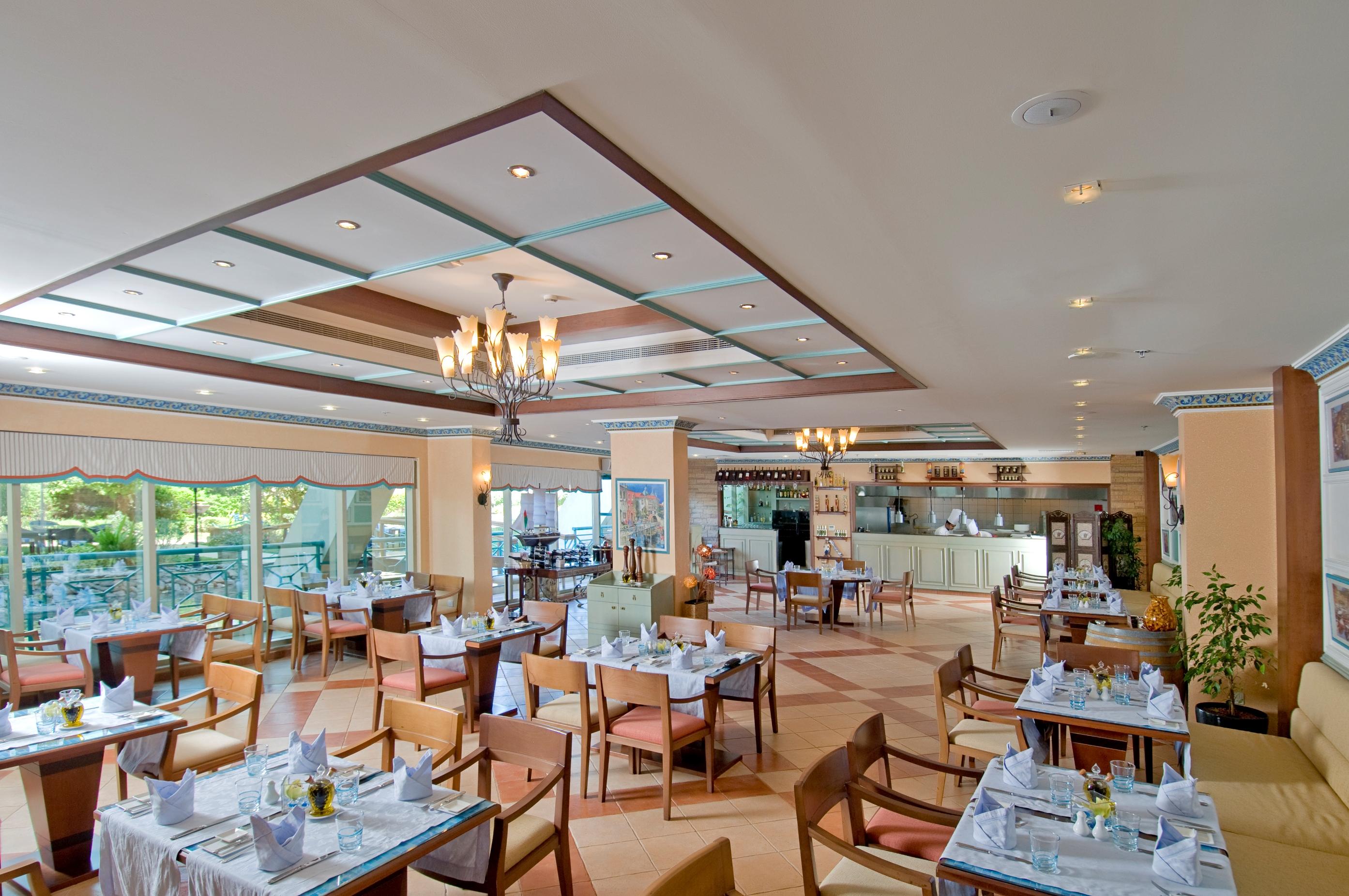 Al Raha Beach Resort & Spa Abu Dhabi Restaurant photo The photo shows the interior of a restaurant. It features a spacious dining area with multiple tables neatly arranged, each set with tableware and cloth napkins. The decor includes a decorative ceiling with lighting fixtures, and large windows that p