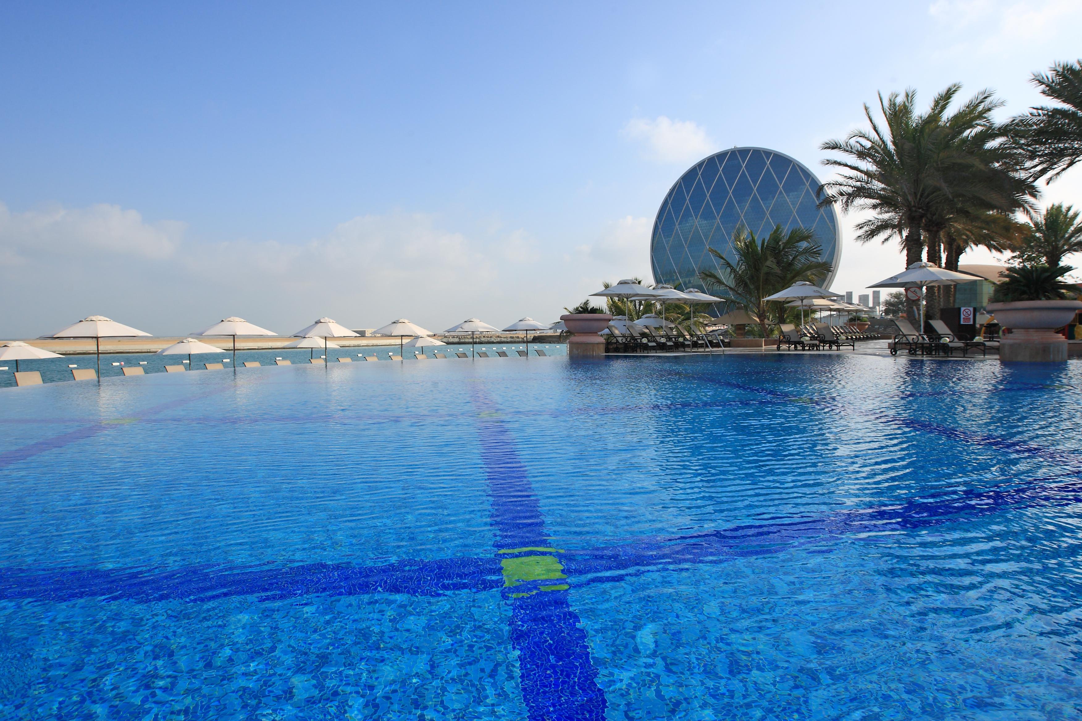 Al Raha Beach Resort & Spa Abu Dhabi Exterior photo The photo features a large, serene swimming pool with clear, blue water reflecting the sky. Surrounding the pool are several lounge chairs and umbrellas, suggesting a relaxed, resort-like atmosphere. In the background, a distinctive building with a r