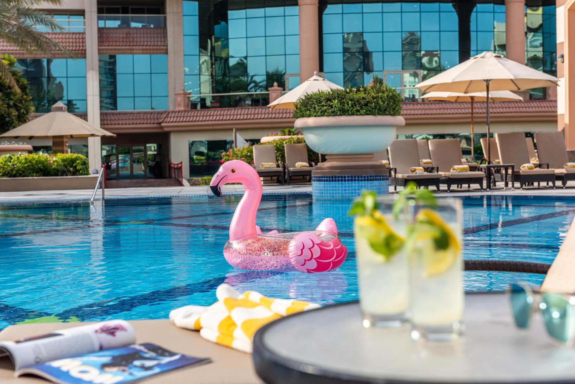 Al Raha Beach Resort & Spa Abu Dhabi Exterior photo The photo depicts a bright and inviting outdoor swimming pool surrounded by a luxurious setting. In the pool, there is a large pink flamingo float, adding a fun and whimsical touch. To the side, a table is set up with two refreshing drinks, likely co