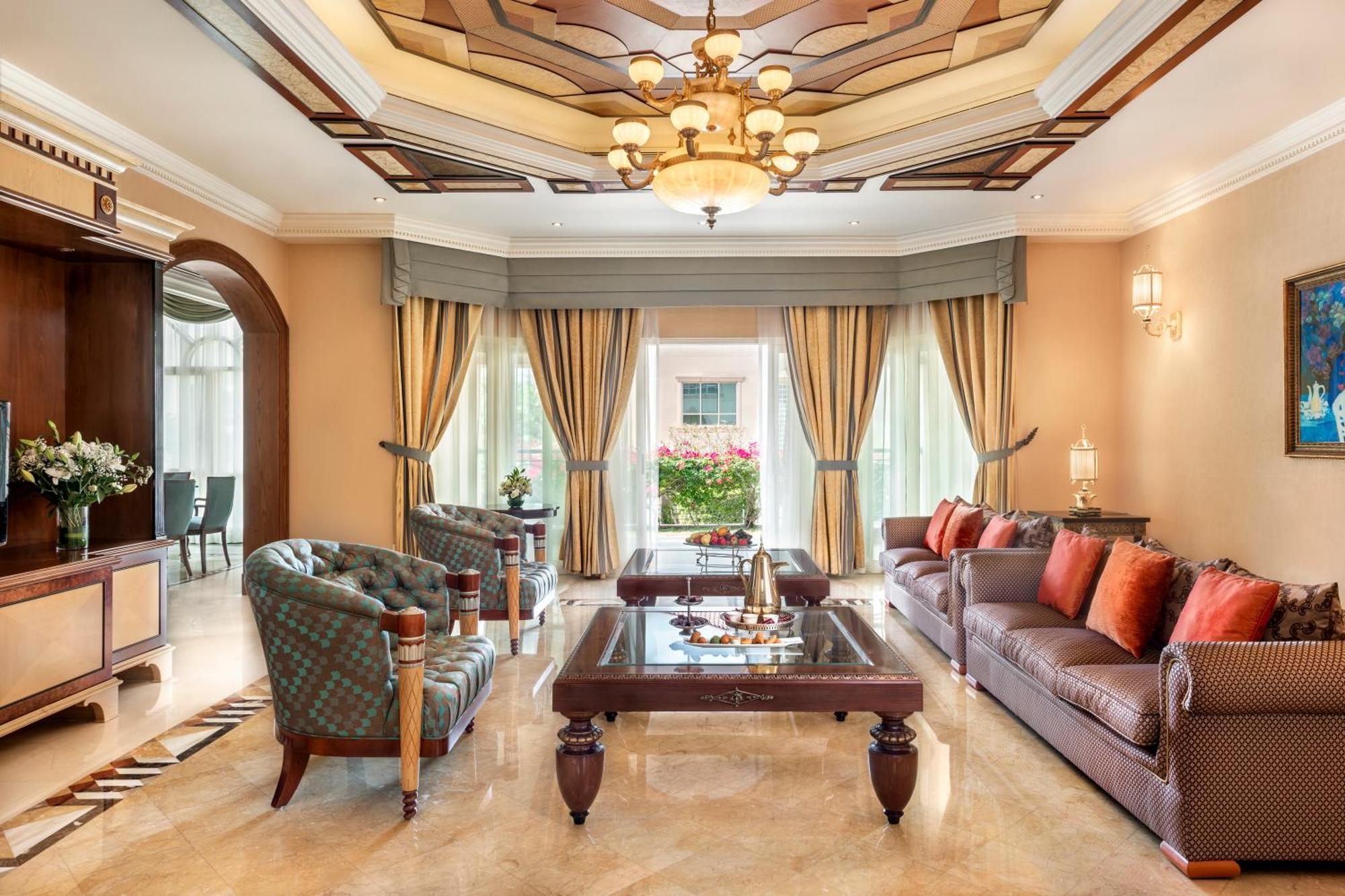 Al Raha Beach Resort & Spa Abu Dhabi Exterior photo The photo shows an elegantly designed living room. It features a high, intricately patterned ceiling with a chandelier hanging in the center. The room is filled with natural light from large windows covered with flowing curtains. 

There are two diff
