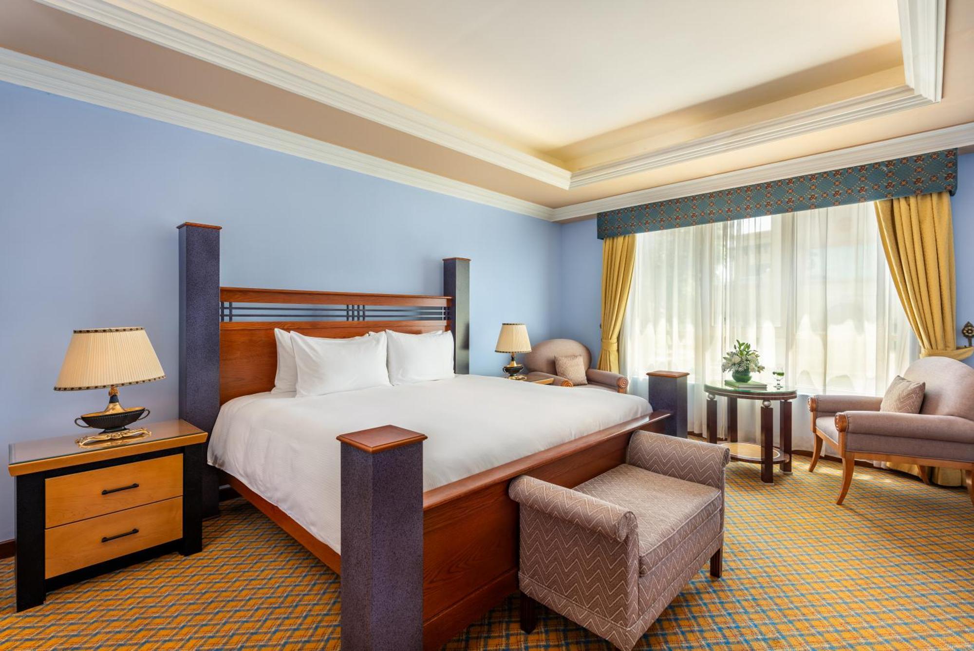 Al Raha Beach Resort & Spa Abu Dhabi Exterior photo The photo shows a well-decorated hotel room. It features a large bed with a wooden frame, neatly made with white bedding. On either side of the bed are bedside tables with lamps. There is a comfortable chair next to a window that has sheer curtains a
