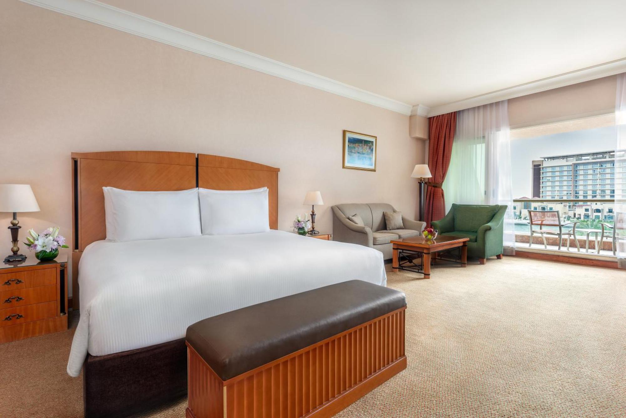 Al Raha Beach Resort & Spa Abu Dhabi Exterior photo The photo shows a well-furnished hotel room featuring a large bed with white linens and a wooden headboard. There's a brown bench at the foot of the bed. To one side, a sitting area includes a sofa and a coffee table, complemented by two lamps. Large