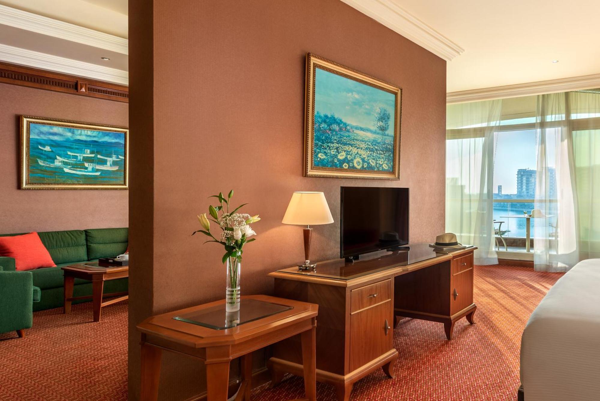 Al Raha Beach Resort & Spa Abu Dhabi Room photo The photo shows a hotel room with a warm, inviting atmosphere. There's a large window that provides natural light and a view, likely overlooking a cityscape or water. On one wall, there's a piece of artwork featuring a landscape, and nearby, a small 