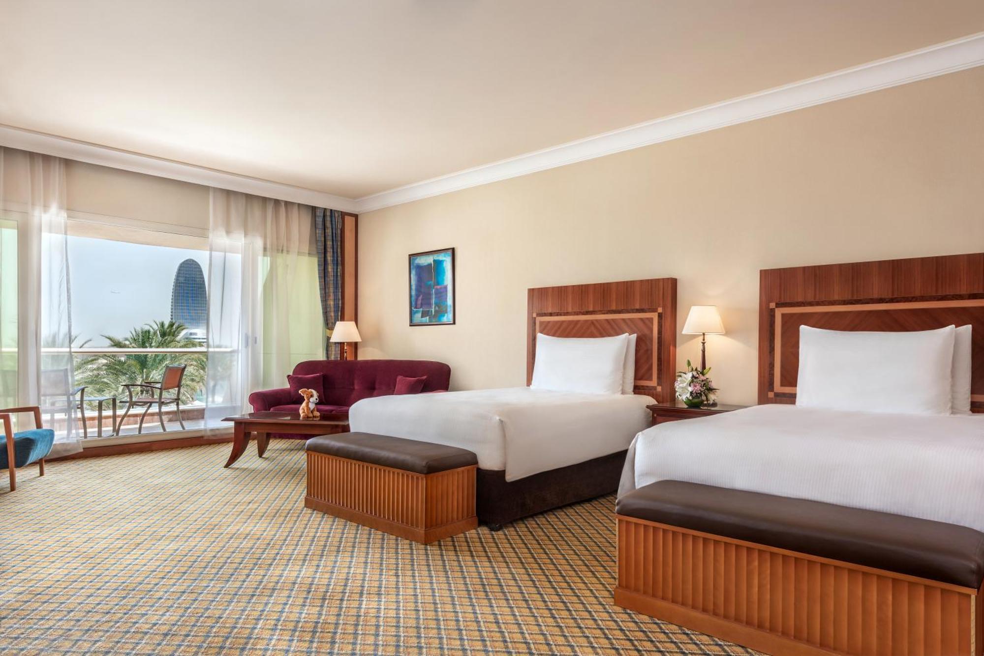Al Raha Beach Resort & Spa Abu Dhabi Room photo The photo shows a hotel room featuring two twin beds with wooden headboards. The room has a light, neutral color scheme, with a large window that provides natural light and a view of the outdoors. There is a small seating area with a purple sofa and 