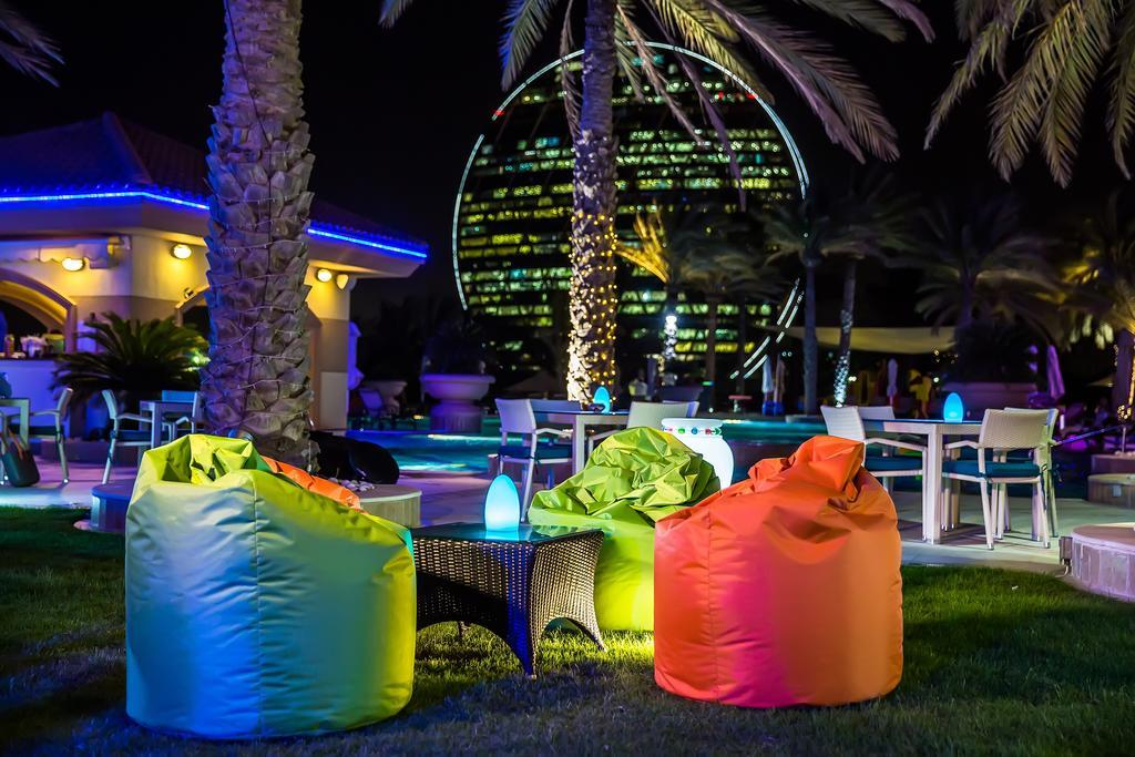 Al Raha Beach Resort & Spa Abu Dhabi Exterior photo The photo shows a vibrant outdoor lounge area at night. In the foreground, there are colorful bean bag chairs—one in bright green, one in yellow, and one in orange. A small table sits between the bean bags. In the background, illuminated palm trees a