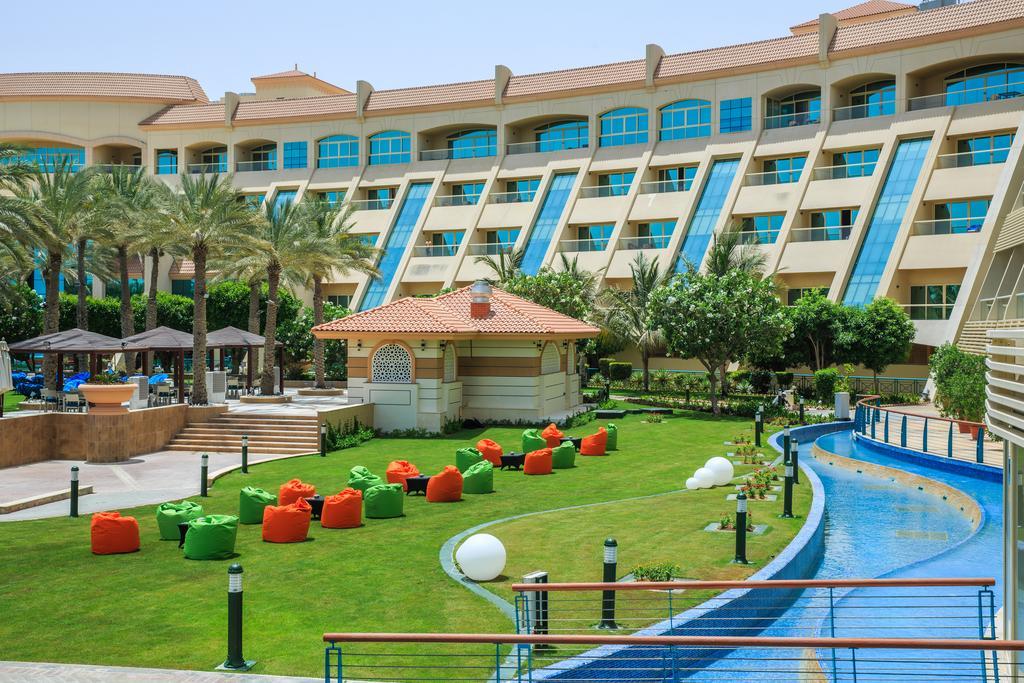 Al Raha Beach Resort & Spa Abu Dhabi Exterior photo The photo depicts a modern hotel complex with multiple stories and large windows. In the foreground, there is a well-maintained lawn featuring colorful lounge chairs in shades of orange and green. A pathway runs alongside a blue water feature, creati