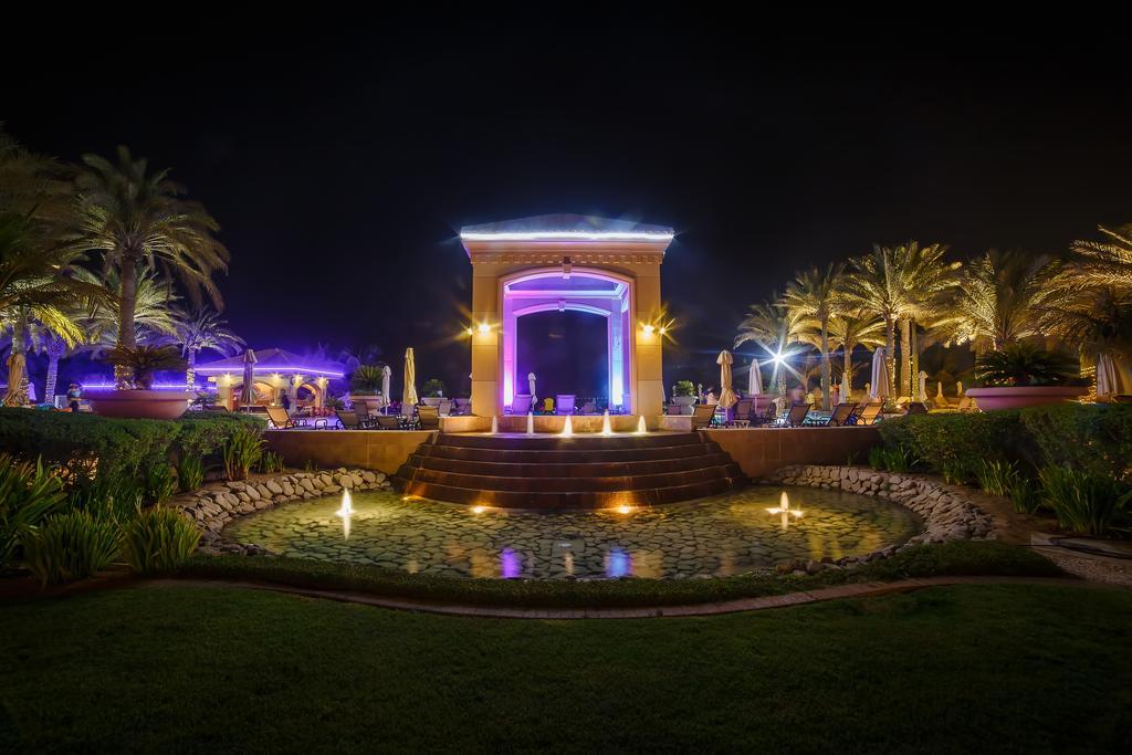 Al Raha Beach Resort & Spa Abu Dhabi Exterior photo The photo depicts a beautifully lit outdoor scene at night. In the foreground, there is a serene water feature with a circular pond, and small fountains creating gentle ripples. Surrounding the pond are lush green lawns and ornamental plants. 

In th