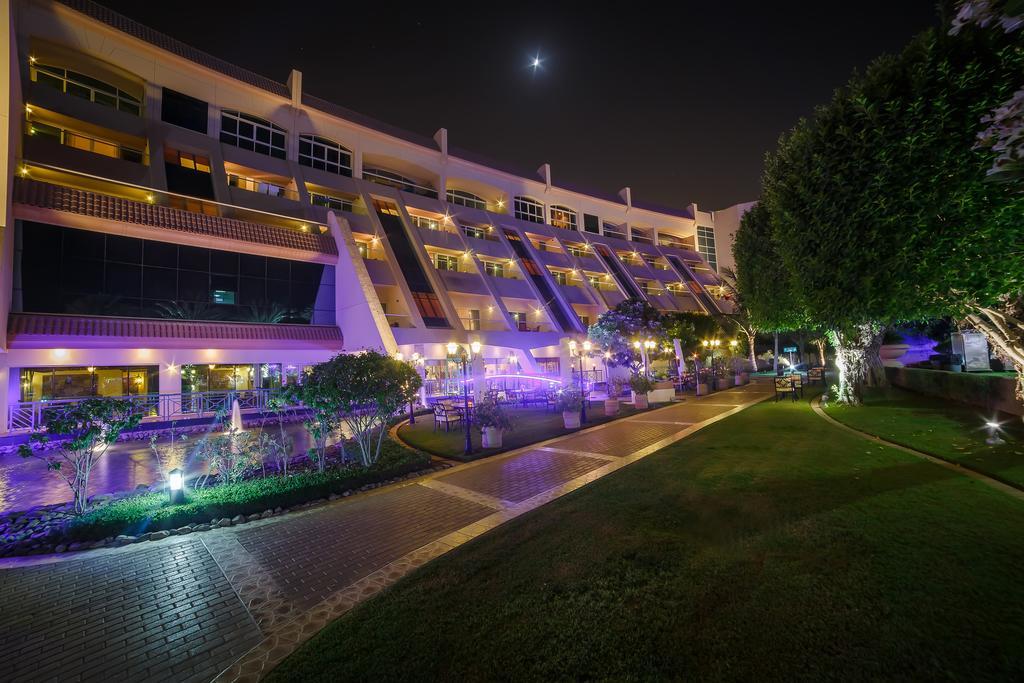 Al Raha Beach Resort & Spa Abu Dhabi Exterior photo The photo shows a modern building illuminated at night, with a vibrant purple hue. The structure has multiple floors, each with balconies overlooking a landscaped area. In the foreground, there is a pathway lined with well-manicured grass and shrubs,