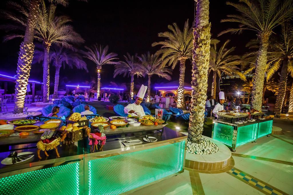 Al Raha Beach Resort & Spa Abu Dhabi Exterior photo The photo depicts an outdoor dining area at night, featuring several chefs in white uniforms and hats working at a food station. The setting is illuminated by colorful lights, with glowing green accents on the serving tables. Surrounding the area are